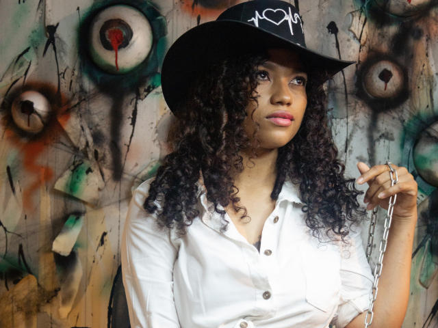 KellyAmbaris with curly black hair, wearing a black hat with a white heart, white button-down shirt, holding a chain, pink lipstick, and ring, looking slightly to the side.