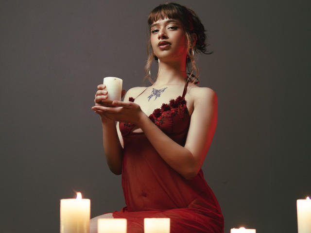 CamiMonet with brown hair in a bun, red lace babydoll, holding a candle, sitting among lit candles, showing a butterfly tattoo on her chest, looking down.