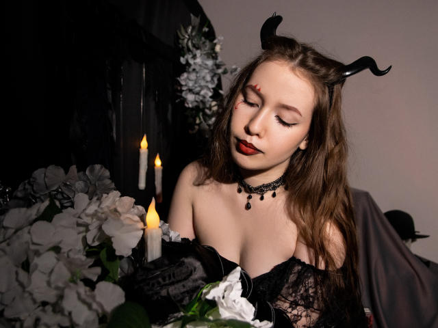  DanielleSweetty with black horns, red dot on forehead, black lace dress, red lipstick, choker, and candles, looking down at white flowers. Her tits are visible, medium-sized.
