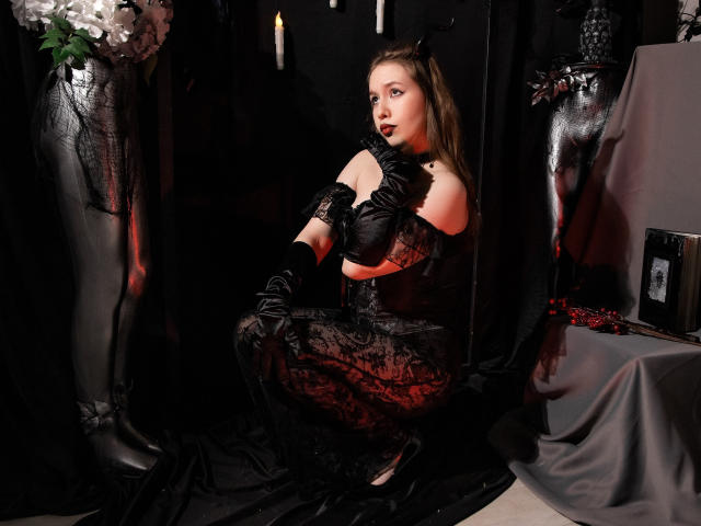  DanielleSweetty sits in a dimly lit, dark room. She wears a black lace dress, matching gloves, and black boots. Her light brown hair is loose, and she has red lipstick. She looks to the right with a serious expression. The background has black curtains, a black vase with white flowers, and a gray cloth-covered table. The lighting is soft, highlighting her pale skin and the textures of her clothing. The overall composition is dark and Gothic.