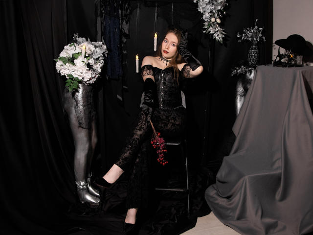  DanielleSweetty in a black lace dress and gloves, sitting on a stool, holding red berries. She has fair skin, red lipstick, and dark brown hair. The background features black curtains, white and black floral arrangements, and a table with a grey cloth. The image has a gothic, Victorian vibe with sharp contrasts and dark, rich textures. Her dress is detailed with intricate lace patterns. The overall composition is balanced, with DanielleSweetty as the central focus.