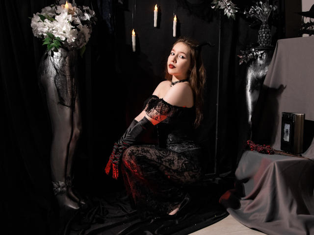 DanielleSweetty trägt ein schwarzes Spitzenkleid mit roten Akzenten, lange schwarze Handschuhe und Teufelshörner, sitzt auf schwarzem Stoff, umgeben von Kerzen und weißen Blumen und wirkt verführerisch..