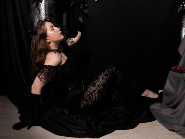 DanielleSweetty, con i capelli castani, un abito di pizzo nero e guanti, seduta su un tessuto nero, con gli occhi chiusi e la bocca aperta, indossa una collana nera, motivi di pizzo nero, scarpe nere e un panno grigio..