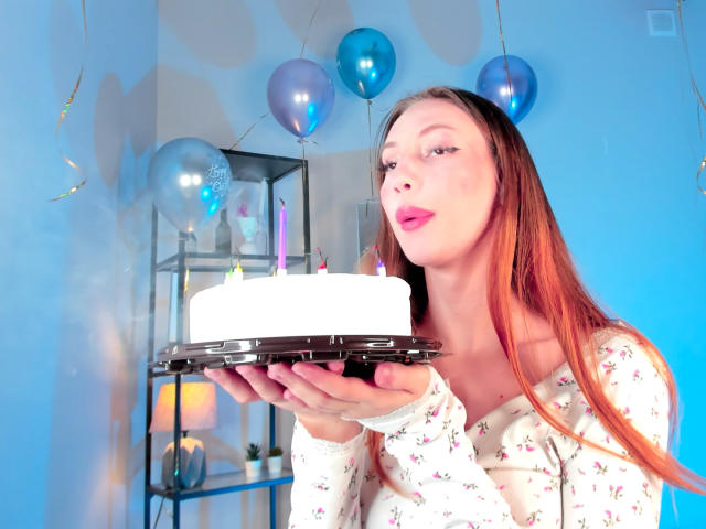ElfinVibes with long red hair, pink lipstick, floral shirt, holding white cake with candles, looking at camera, blue balloons in background.