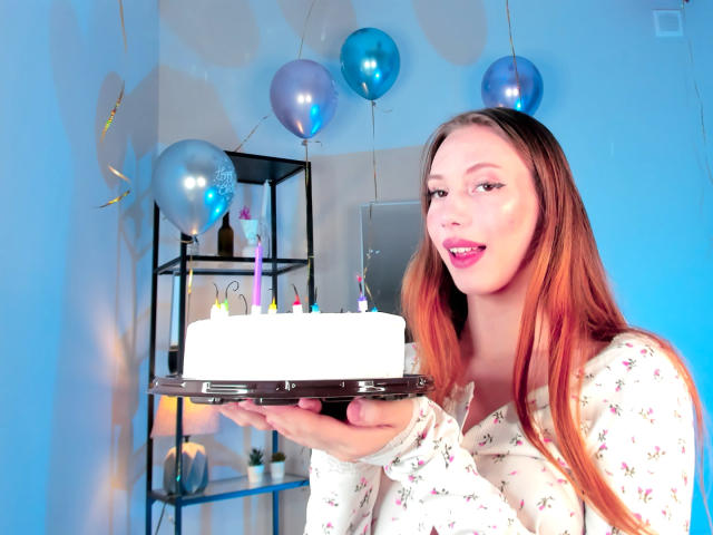 ElfinVibes com cabelos vermelhos longos, vestindo uma camisa florida, segurando um bolo branco com velas coloridas, sorrindo, balões azuis ao fundo, em pé, batom rosa, unhas rosa..
