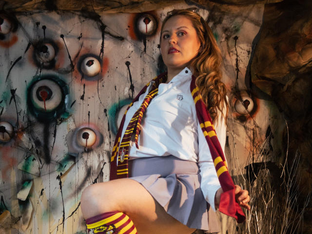 Lauren Spitz mit langem, blondem Haar, die ein weißes Hemd, einen grauen Rock und einen rot-gelb gestreiften Schal sowie knielange Socken trägt, lehnt sich an eine mit Graffiti beschmückte Wand, auf der Augen gemalt sind..