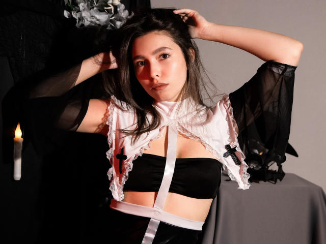 LounaLove with dark hair, wearing a black and white sheer top, exposing her tits, arms raised, looking at camera, black and white background, candle on left.