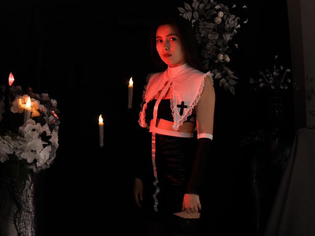 LounaLove in a black and white maid outfit, exposing small tits with black crosses, standing in dim red light, arms down, with candles and white flowers on the left.