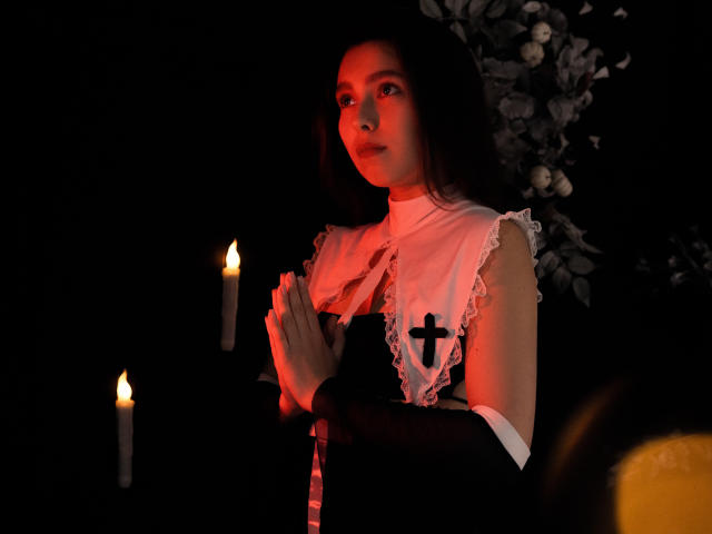 LounaLove with dark hair, wearing a black and white cross-adorned dress, praying with hands clasped, candlelight glowing on her face, in a dimly lit, dark setting.