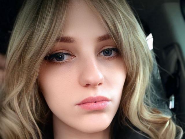 JoseFairy has blonde wavy hair, blue eyes, and pinkish lips. Her face is centered with a neutral expression, wearing a black top. Her tits are not visible.