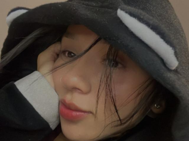 KloeThompson's face, wearing a black panda hood with white eyes, has her hand under the hood, black hair covering her eye, slightly smiling, with red lips.