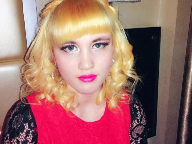 SophieDivyne heeft blond krullend haar, felroze lippenstift en blauwe ogen. Ze draagt een rode kanten jurk met zwarte kanten mouwen. Haar borsten zijn bedekt door de jurk..