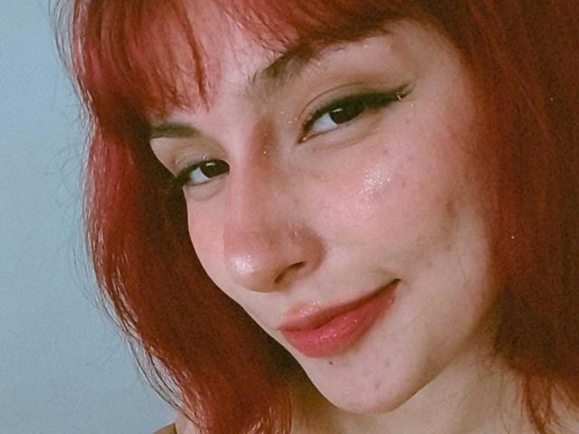 IsabellaHallad has red hair, a cute nose piercing, and a slight smile with pink lips. Her skin is shiny with some small blemishes. She has black eyeliner.