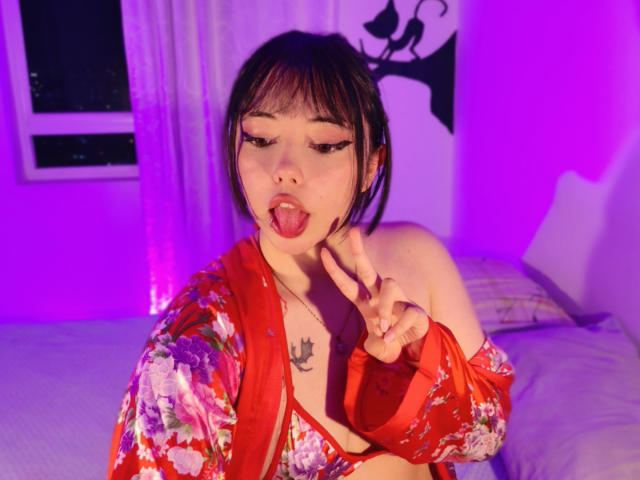 Arieyaara with bangs, black hair, and a nose ring, sticking out her tongue, wearing a red floral robe, flashing a peace sign, and exposing her tits in a purple-lit room.