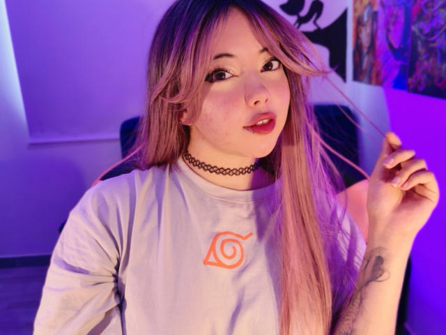 Arieyaara, avec ses cheveux blond-rosés, son collier et sa chemise blanche, tenant une mèche de cheveux, montrant un tatouage sur son bras droit, éclairage violet-bleu, et un léger sourire, avec du rouge à lèvres rouge..