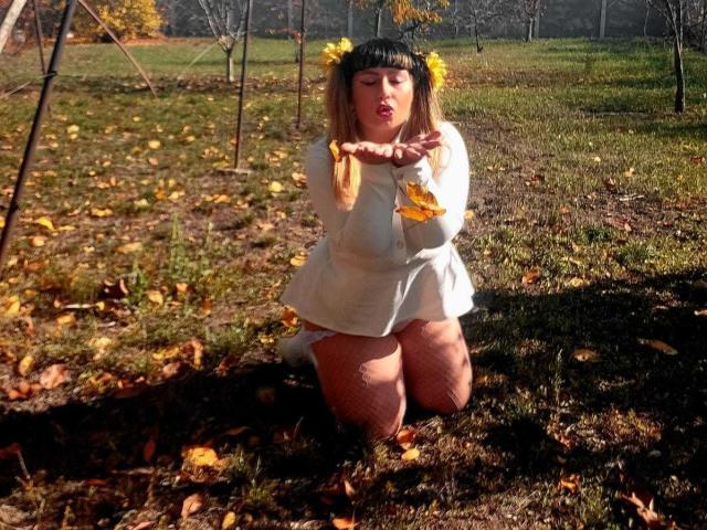 MirayDesire kneels on a grassy field, wearing a white, long-sleeve dress with yellow flowers in her straight, black-fringed hair. Her eyes are closed, and she has her right hand out, fingers spread. The sunlight casts shadows on the ground, and yellow leaves scatter around her. She's in a natural setting with leafless trees in the background. Her dress contrasts with the brown grass and fallen leaves. The image has a warm, autumnal feel.