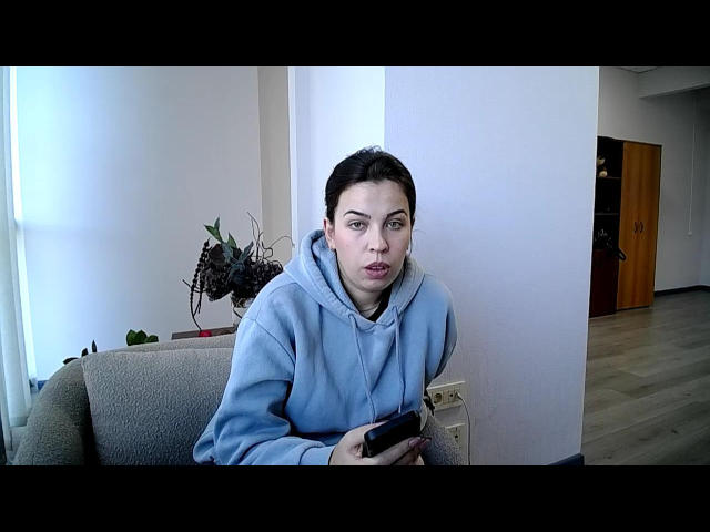 Glariya with short dark hair, wearing a light blue hoodie, holding a black remote, sitting on a beige couch, looking surprised with slightly parted lips.
