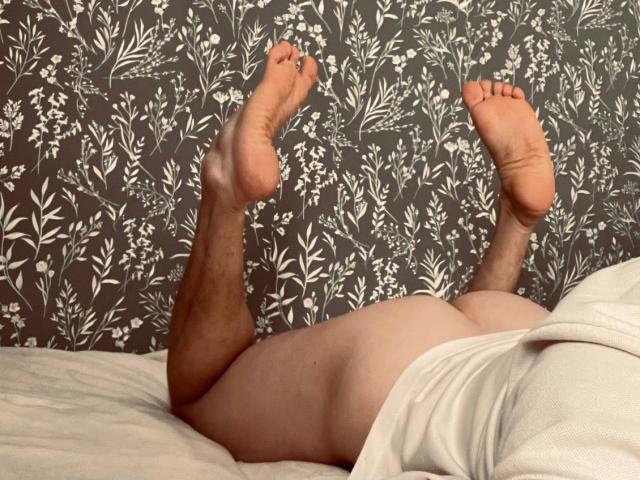 LuckyFlame's ass is up in the air, legs bent at the knees, feet pointed upward, lying on a bed with a white blanket, showing their bare back and buttocks.