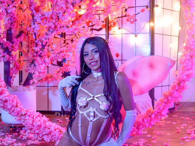 ClarisCampbell with long black hair, wearing a pink strappy lingerie set, white gloves, fairy wings, and floral pasties covering her tits, surrounded by pink cherry blossom petals.