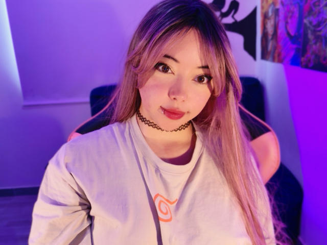 Arieyaara with long pinkish-blonde hair, wearing a white shirt with an orange spiral, black choker, and nose ring, sitting in purple-lit room, looking at camera.