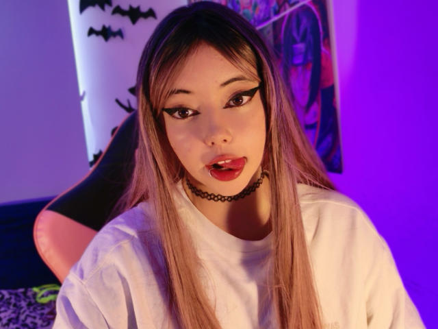 Arieyaara with long brown hair, black eyeliner, red lipstick, choker, and white shirt, tongue slightly out, purple and blue lighting, looking at camera, legs in background.