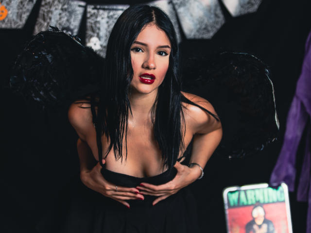 LizzeJoy with black wings, red lipstick, and black dress, hands on her tits, looking up with dark eyes, long black hair, and a sexy, goth vibe.