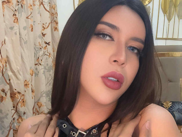 GoddessCeline with dark brown hair, full pink lips, and black leather collar, hands on her chest, looking up with half-closed eyes, soft light on her face.