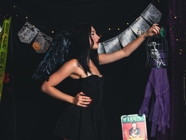 LizzeJoy with black wings, red lipstick, and black dress, reaches for a helmet with her right hand, showing off her tits. Purple jacket and poster in the background.