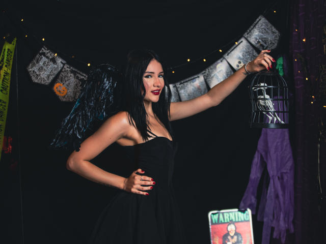 LizzJoy with black wings, wearing a strapless black dress, raises a birdcage with one hand, showing off red-painted nails, and has a sexy, confident look.