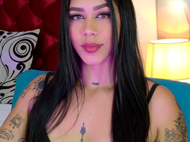 AgathaVelez69 has long black hair, purple lipstick, tattoos on her arms and chest, and is wearing a black top, sitting with a lamp and patterned pillow in the background.