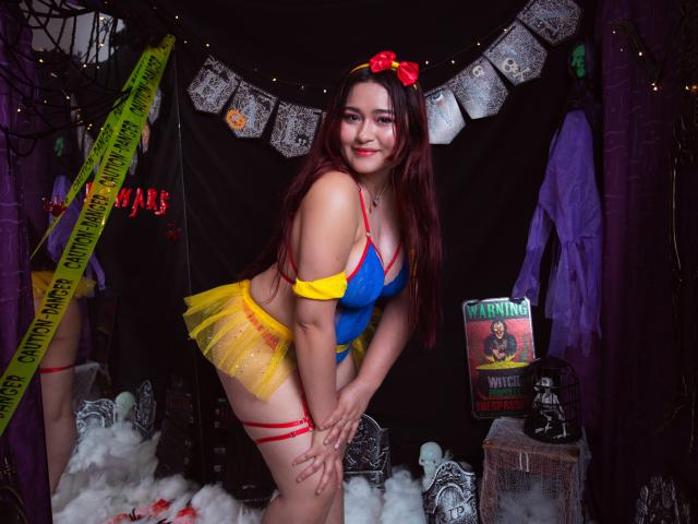 MaddieMoon con lunghi capelli rossi, costume blu e giallo, spalline rosse, gonna gialla, fiocco, sorridente, piegata in avanti, che mostra i suoi piccoli seni, in un contesto a tema Halloween..