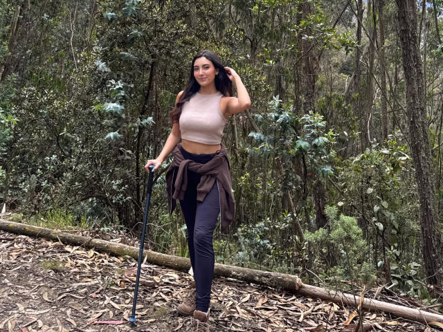 CarlaFranco has long dark hair, wearing a pink crop top, black pants, brown jacket tied around waist, holding a blue walking stick, standing in a forest with a log.