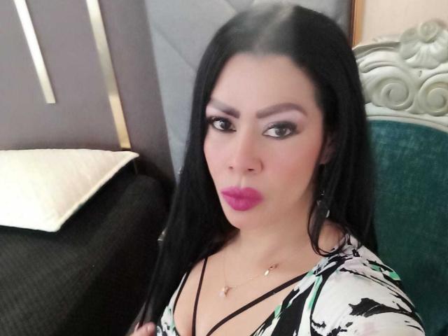 SofiMontes, avec ses longs cheveux noirs, son rouge à lèvres rose et son haut fleuri, faisant la moue, montrant sa poitrine, assise sur une chaise verte, regardant l'appareil photo..
