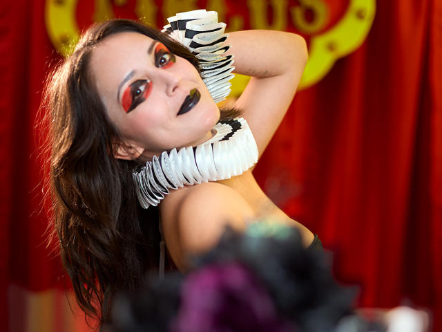 MollyMarmalade con un trucco scuro, ombretto rosso, rossetto nero, indossa una collana bianca con volant e guanti a righe zebrate, in posa con le braccia alzate, mostrando il seno, davanti a una tenda rossa..