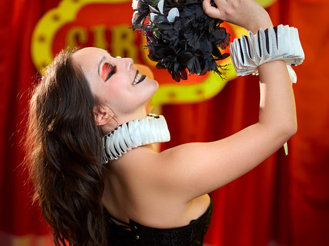 Molly Marmalade, avec ses cheveux noirs, souriante, tenant des plumes noires, portant des poignets blancs froncés et un corset noir, montrant ses dents, sur fond de rideau rouge. Ses seins sont visibles..