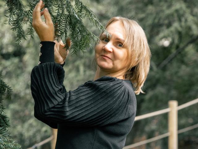 LeilaColleen, con cabello rubio, una camiseta negra con textura y una sonrisa sutil, tocando ramas de pino verdes con ambas manos, de pie al aire libre, mirando ligeramente hacia atrás..