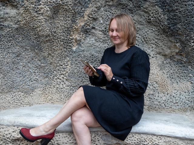 LeilaColleen, with a black dress and red shoes, sits on a stone bench, legs crossed, holding a phone, smiling, and looking at it with a slight tilt.