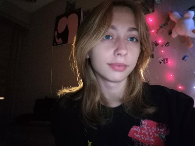 TracyLeex has light brown hair, blue eyes, and pink lips, wearing a black shirt with a red graphic, in dimly lit room with pink fairy lights.
