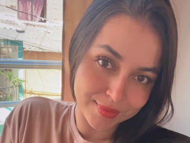 HikariChloe with medium-length black hair, brown eyes, red lipstick, and a cute smile, wearing a pink shirt, showing her soft, fair-skinned face and subtle makeup.