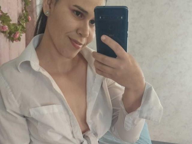 DanTaylor, com um leve sorriso, segurando um telefone, vestindo uma camisa branca aberta, expondo seus seios, em um selfie no espelho. Seu cabelo castanho está preso em um rabo de cavalo..