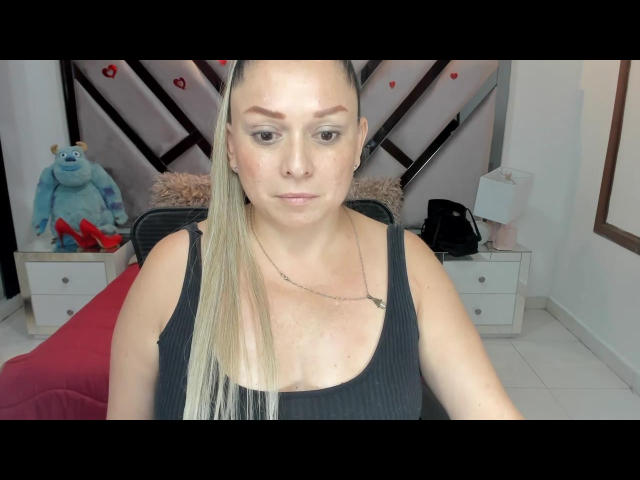Natalia69s with blonde ponytail, black tank top, small tits, and a neutral face, sitting in front of a white wall with a blue plush toy and red heels.