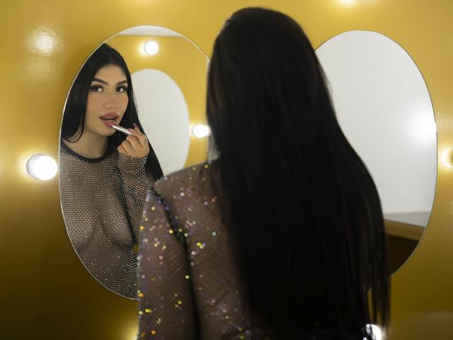 CamilaVerone with long black hair, wearing a sheer, glittery top, looking in a mirror while applying lipstick, showing her boobs and reflection, yellow wall in background.