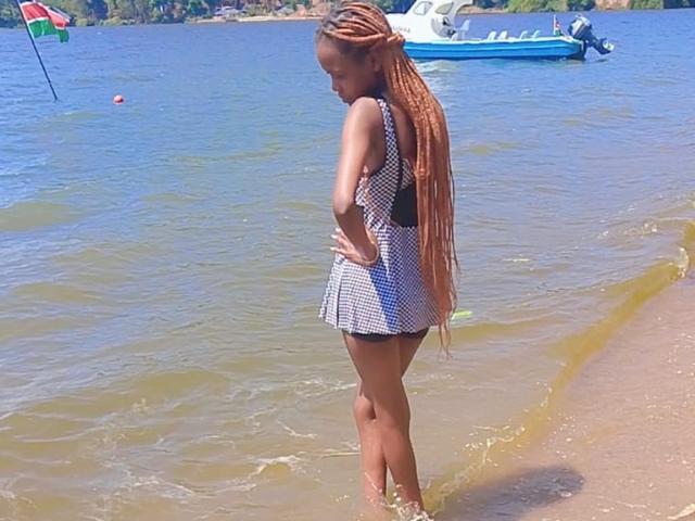 HotyBarbarha with long, orange, braided hair, wearing a black-and-white checkered dress, standing in shallow water with a blue boat and green flag in the background.