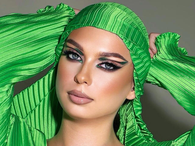 WildHoneyy has green, pleated headwear, blue eyes with thick black eyeliner, and glossy pink lips. She's holding the headwear with both hands, looking straight at the camera.
