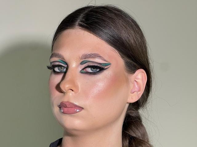 WildHoneyy has dark brown hair, teal winged eyeliner, black eyelashes, glossy pink lips, and a shiny face with a neutral expression. She's looking slightly to the side.