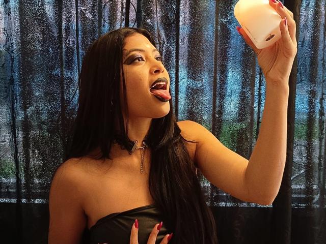 MariaInLove with long black hair, tongue out, holding a pink iPhone, wearing a black choker and strapless top, red nails, showing her tits, against a dark curtain.