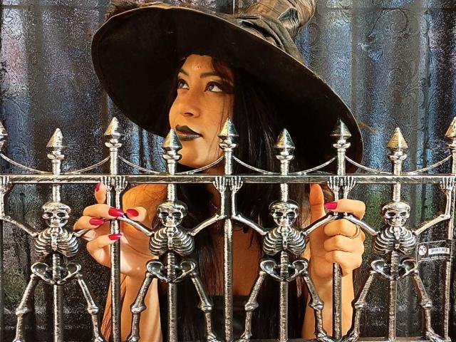 MariaInLove with dark lipstick, black hat, and red nails holding a silver fence with skull decorations, looking to the side. Her hands grip the fence, showing off her nails.