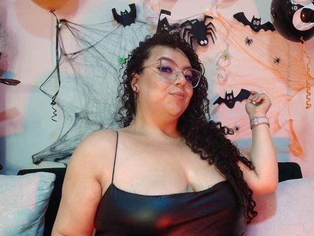 NatashaAmbeer, with curly dark hair and glasses, wearing a black spaghetti-strap top, showing her big tits, and a pink wristband, holding a bat decoration.