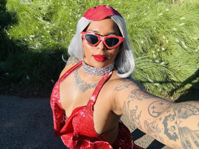 Chili tiene labios rojos brillantes, peluca blanca, vestido rojo, tatuajes en los brazos, gafas de sol rojas, collar de gargantilla, tomando selfie al aire libre.
