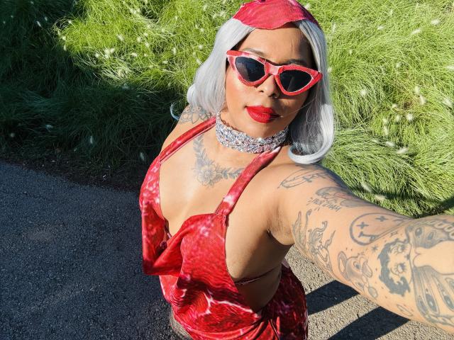 Chili with white hair, red eyes, red lipstick, red dress, red glasses, silver necklace, tattoos, outdoor, sunny, grass background.